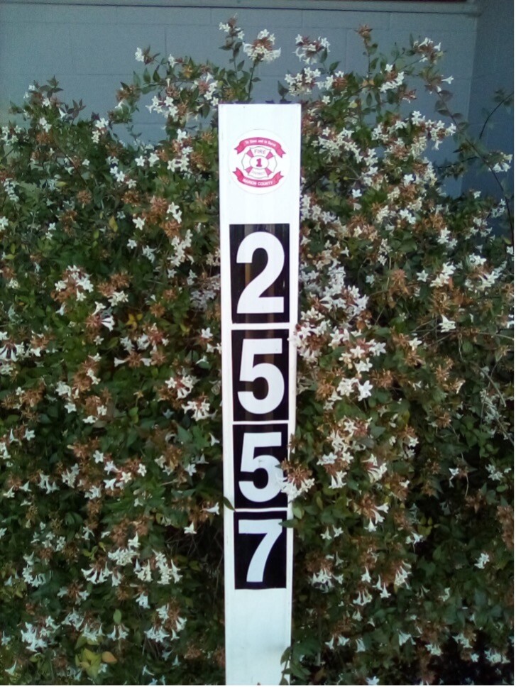 A white post with black numbers "2557" and a red and white logo stands in front of a bush with small white flowers.