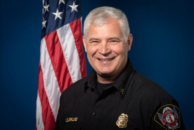 Smiling person in a black uniform with fire department patches stands in front of an American flag against a blue background.