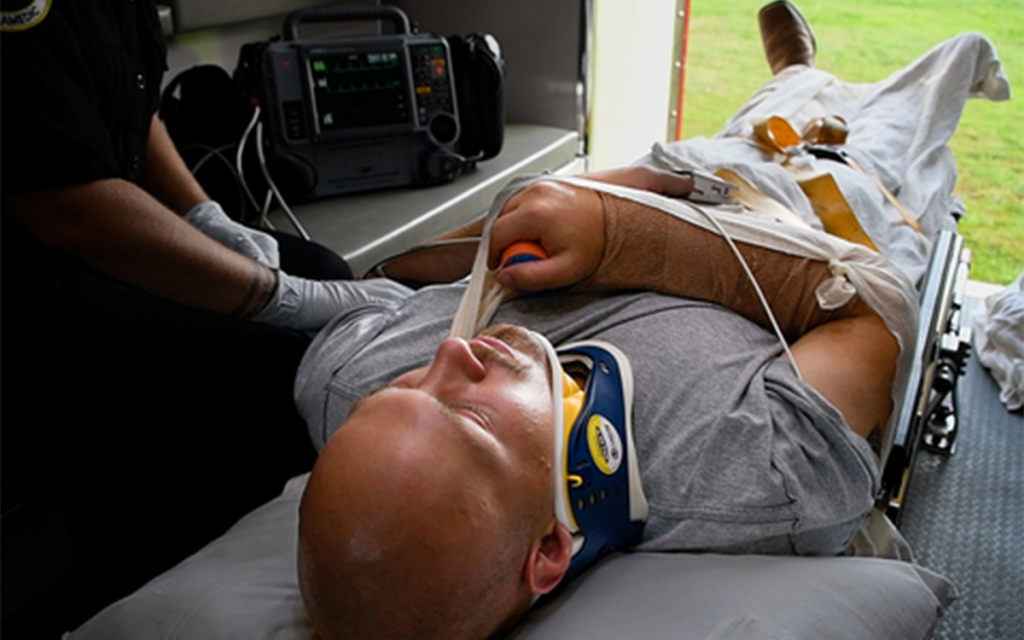 A person with a shaved head is lying on a stretcher in an ambulance, secured with straps and wearing a neck brace. They have a bandaged arm resting on their chest. Medical equipment is visible nearby through the open ambulance door.