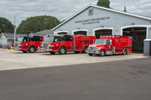 Three red emergency vehicles are parked outside a fire station labeled "Marion County Fire District #1 Four Corners Station." The sky is overcast, and trees are visible in the background.