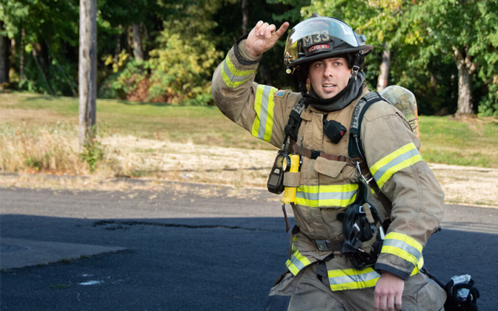 A firefighter in full gear, including a helmet labeled "M53," is kneeling on a road near a grassy area, gesturing upwards with one hand. He appears alert and ready for action, surrounded by trees in the background.