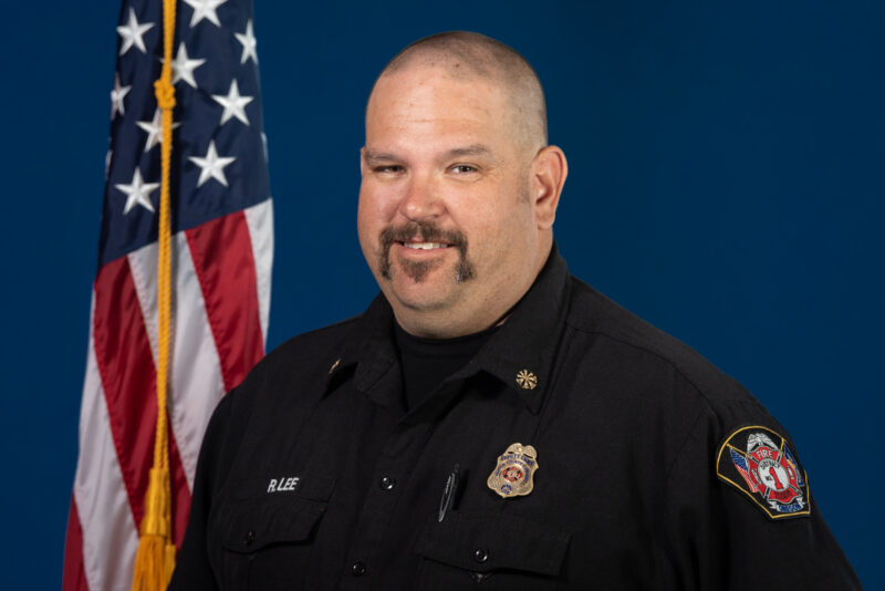 A man wearing a black uniform with badges stands in front of an American flag. The background is a solid blue color.