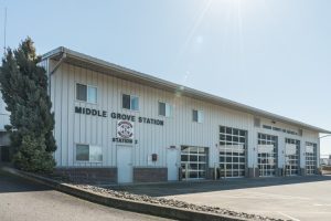 Exterior of a white metal building labeled "Middle Grove Station, Station 2" with signage for "Marion County Fire District #1". The building features large garage doors and windows, with a paved driveway and a tall tree on the left.