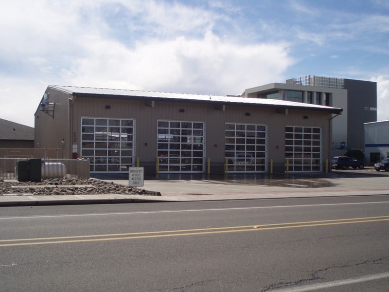 Fire station with four glass-paneled garage doors, situated next to a road. A few cars are parked nearby, and a sign reads "Authorized Vehicles Only." The sky is partly cloudy, and a modern building is visible in the background.