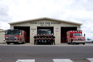 A group of uniformed firefighters stands in front of Clear Lake Fire Station, Engine Co. 725. Two fire trucks and an ambulance are parked on either side of the group. The building is beige with open bays.