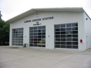 Exterior of a fire station labeled "Labish Center Station, Station 7." The building is white with three garage doors and a paved driveway in front. A few trees are visible to the side.