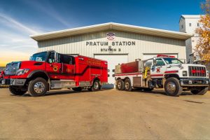 Two fire trucks are parked in front of Pratum Fire Station 3. The station is a white building with a gray roof. The truck on the left is red, and the one on the right is white and red. A blue sky is visible in the background.