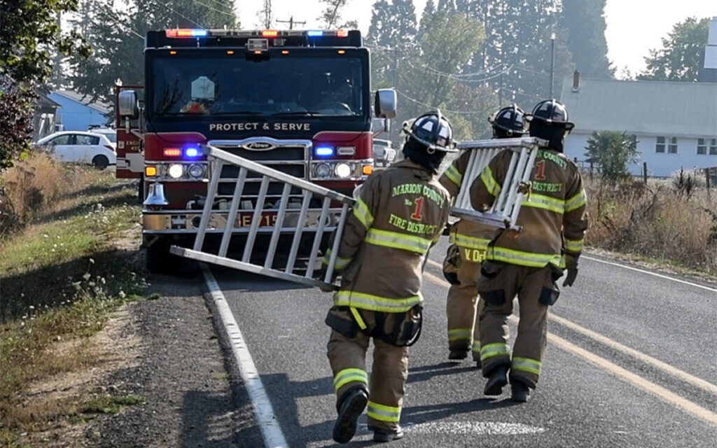 Three firefighters carry ladders towards a red fire truck parked on a rural road. They are wearing protective gear, including helmets and reflective uniforms. Trees and a few buildings are visible in the background.