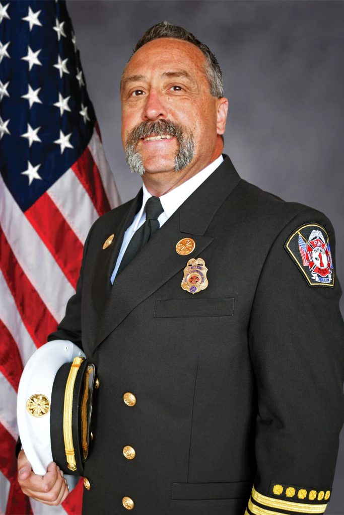 A man in a dark military-style uniform with gold buttons and badges poses holding a white cap. He has a mustache and stands in front of an American flag against a grey background.